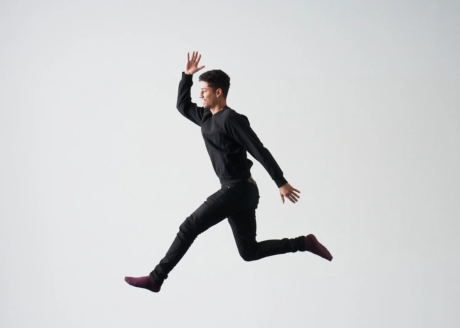 Josh Henderson-Jardim mid-leap in a studio, black sweater and jeans, oxblood socks